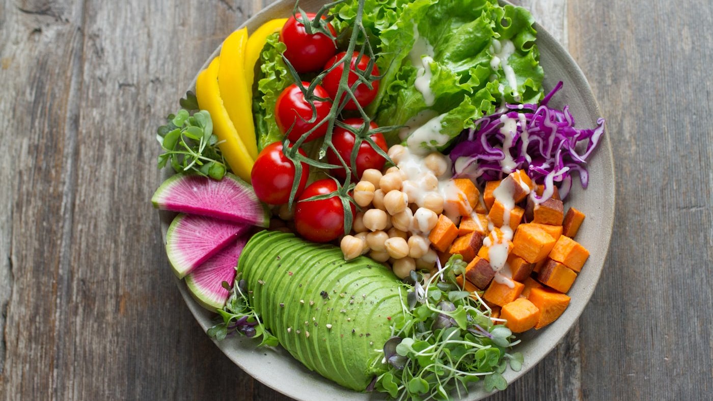 A bowl of vegetables, greens, and whole foods on a plain surface